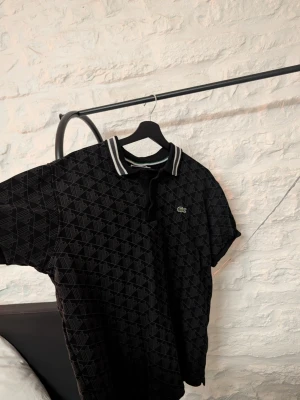 Svart pikétröja från Lacoste - Säljer en svart pikétröja från Lacoste med diskret geometriskt mönster och klassisk krage med vita ränder. Tröjan har korta ärmar och den ikoniska gröna krokodilloggan på bröstet. Perfekt för en stilren och sportig look.