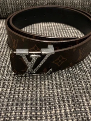 Brunt bälte  - Säljer ett brunt bälte med klassiskt monogrammönster och en silverfärgad spänne. Skärpet är tillverkat i skinn och har en elegant design som sticker ut. Perfekt för dig som vill ha en lyxig accessoar med ikonisk stil.