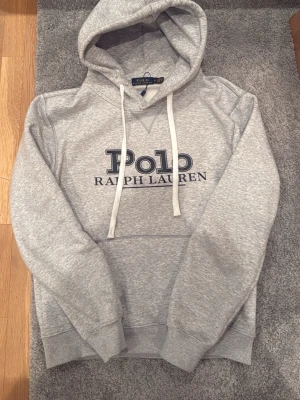  Polo Ralph Lauren Hoodie  - Snygg grå hoodie från Polo Ralph Lauren med stor logga framtill. Klassisk modell med huva och vita dragsnören, samt ribbade muddar vid ärmslut och nederkant. Perfekt för en chill och stilren look.