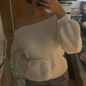 Vit off shoulder stickad tröja - Trendig vit stickad tröja med  an off shoulder sopad ringning och breda ribbade muddar. Tröjan har`` avslappnad passform och lå sopade axlar, perfekt för en chill och snygg look. Passar till jeans eller kjol för en casual stil.