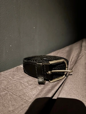 Svart bälte med metallspänne - Ett svart smalt bälte med ett klassiskt metallspänne. Bältet har enkel design och passar perfekt till jeans. Materialet är troligtvis syntet och spännet är i silverfärgad metall. Perfekt för att ge din outfit en stilren touch.