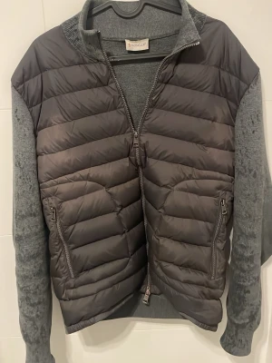 Grå pufferjacka från Moncler - Snygg grå pufferjacka från Moncler med stickade ärmar och rygg i mjukt material. Framsidan är quiltad med dragkedja och två fickor med dragkedja. Perfekt för kalla vinterdagar och har en modern, sportig look. Och om ni undrar varför den är fläckig är de bara vatten!