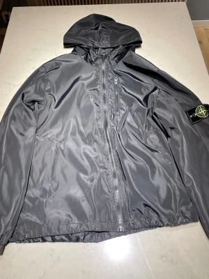 Svart vindjacka från Stone Island - Snygg svart vindjacka från Stone Island med huva och dragkedja framtill. Jackan har en ikonisk patch på vänster ärm och är tillverkad i ett glansigt, lätt material som står emot vind. Perfekt för dig som gillar streetwear och vill ha en stilren look.