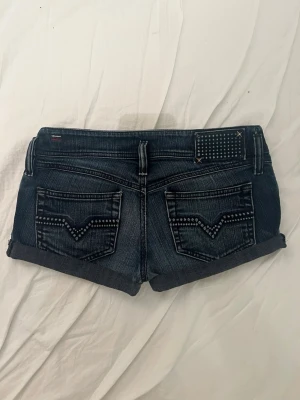 Mörkblå jeansshorts från Diesel - Snygga mörkblå jeansshorts från Diesel med uppvikta benslut och dekorativa prickar på bakfickorna.  Midjemåttet rakt över: 39cm, längden på shortsen: 26cm. Står ej storlek i shortsen, men ja upplever det som en medium