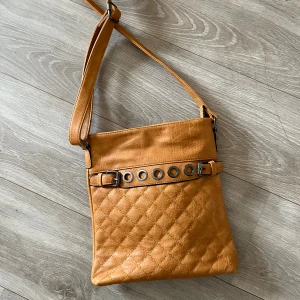 Karamellbrun axelväska med nitar - Snygg karamellbrun axelväska i quiltad konstläder med justerbart axelband. Framsidan har dekorativt bälte med metallringar och nitar. Baksidan har en liten dragkedjeficka. Ny med prislapp 