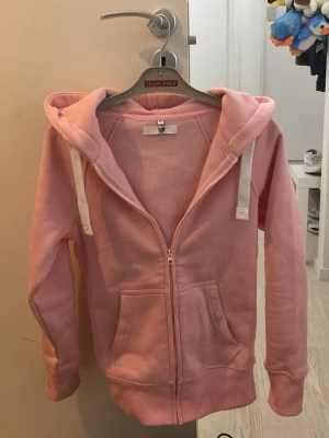 Ljusrosa zip hoodie XS - Ljusrosa hoodie med dragkedja från Resteröds. Klassisk modell med huva och vita dragsnören, samt två stora fickor framtill. Tillverkad i mjukt sweatshirtmaterial som är perfekt för chill dagar. Ribbstickade muddar vid ärmslut och nederkant.
