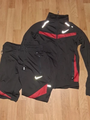 Nike svart träningsset med röda detaljer - Snyggt svart träningsset från Nike med röda detaljer och vita reflexloggor. Setet består av en zip-jacka med hög krage och två fickor samt matchande byxor med dragsko och fickor. Perfekt för träning eller chill. Materialet är mjukt och stretchigt.
