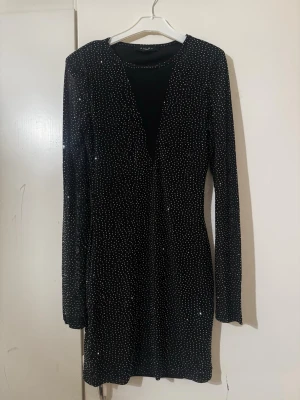 Svart glittrig kort klänning - Säljer en svart kort klänning från New yorker med glittriga detaljer och djup svart/genomskinlig v-ringning. Klänningen har lång ärm och en tight passform som framhäver figuren. Perfekt för dig som vill sticka ut med en elegant och trendig look.