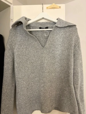 Grå stickad tröja med krage - Säljer en stilren grå stickad tröja från Only med bred krage och v-ringning. Tröjan har långa ärmar och ribbstickade muddar vid ärmslut och nederkant. Perfekt för lager på lager och en avslappnad look.