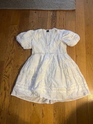 Vit kort klänning med puffärm - Supersöt vit klänning med puffärmar, v-ringning och knappar framtill. Klänningen har spetsdetaljer och är gjord i ett lätt bomullstyg. Perfekt för varma dagar och har en romantisk vibe. 