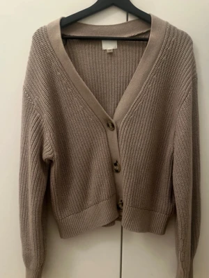 Beige stickad kofta med knappar - Mysig beige stickad kofta med v-ringning och stora knappar framtill. Koftan har ribbade muddar vid ärmslut och nederkant samt en avslappnad passform. Perfekt att slänga över en t-shirt för en chill look.