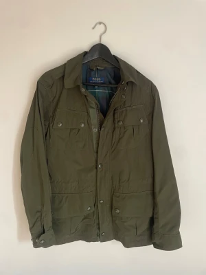 Polo Ralph Lauren jacka  - Stilren olivgrön jacka från Polo Ralph Lauren med rutigt foder i blått och grönt. Jackan har flera fickor framtill, knappar och dragkedja. Storlek XS. Jackan är i princip nyskick, nypris ca 2000 kr