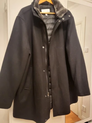 Svart kappa från Calvin Klein XXL - Stilren svart kappa från Calvin Klein i storlek XXL. Kappan har en mjuk fuskpäls vid kragen, dragkedja och knappar framtill samt två sidofickor. Insidan är quiltad för extra värme och komfort. Perfekt för kalla dagar med en klassisk look.