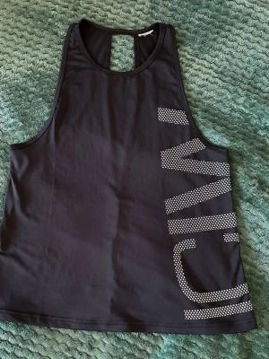 Svart ICIW  linnetopp med tryck - Sportig svart linnetopp från ICIW  med stort vitt logotyptryck längs sidan. Toppen är ärmlös och har en öppen detalj i ryggen samt en lös passform. Perfekt för träning och tillverkad i ett lätt, syntetiskt material som andas.