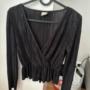 Svart fest topp från HM - Snygg svart omlottopp från Gina Tricot med plisserat tyg och lång ärm. Toppen har djup v-ringning och volang nertill.