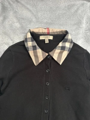 Svart skjorta med rutig krage Burberry - Säljer en svart skjorta från Burberry med klassisk rutig krage i beige, svart och grått. Skjortan har knappar framtill och lång ärm. Ikonisk Burberry-logga broderad på bröstet. Perfekt för dig som gillar stilrena detaljer.