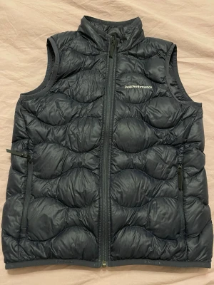 Marinblå dunväst Peak Performance - Snygg marinblå dunväst från Peak Performance med krage och två dragkedjeförsedda fickor. Västen har quiltat mönster och Peak Performance-logga på bröstet. Perfekt för lager-på-lager och håller dig varm under kyliga dagar.