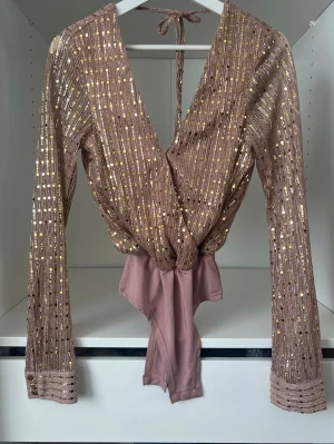 Rosa glittrig omlottbody med paljetter - Supersnygg rosa omlottbody med långa ärmar och guldiga paljettdetaljer. Djup v-ringning och knytband i nacken ger en trendig look. Stretchigt material som sitter bekvämt och markerar midjan. Perfekt för dig som vill sticka ut lite extra.
