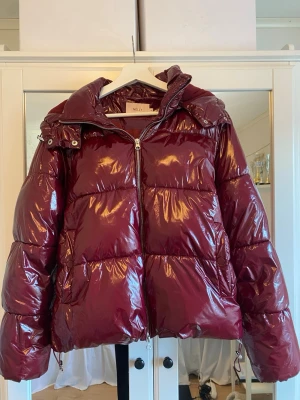 Vinröd pufferjacka Nelly - Snygg slutsåld vinröd pufferjacka från Nelly med glansig finish, dragkedja framtill och hög krage. Perfekta vinterjackan. Fint skick bortsett från att resåren i nederkant har gått av inifrån, har löst detta provisoriskt med en säkerhetsnål, går att laga bättre. Se bild 4.