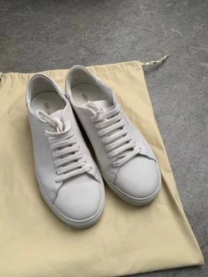 Axel Arigato clean 90s - Snygga vita sneakers från Axel Arigato i stilren design. Skorna är låga och gjorda i slätt läder med vita skosnören och diskret guldfärgad logga på sidan. Perfekta för en clean och minimalistisk look. 