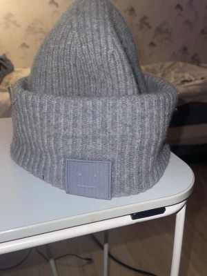 Grå ribbstickad mössa från Acne Studios - Säljer en grå ribbstickad mössa från Acne Studios med den klassiska face-loggan framtill. Mössan är i mjukt och varmt material, perfekt för kyliga dagar. Den har en bred uppvikt kant och en stilren, minimalistisk look.