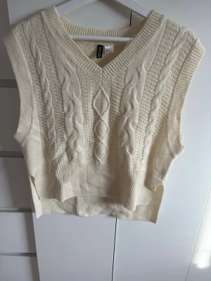 Vit stickad väst med kabelmönster - Vit kabelstickad väst från Divided med V-ringning och bred ribbad nederkant. Västen är ärmlös och har snygga flätade detaljer framtill. Materialet är mjukt och passar perfekt för lager-på-lager.  Frakt betalar man själv