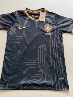 Svart Brasilien Nike matchtröja - Snygg svart matchtröja från Nike med Brasilien-emblem och gul Nike-logga på bröstet. Tröjan har guldfärgade detaljer och grafiskt mönster framtill, samt rund halsringning och korta ärmar. Perfekt för fotboll eller träning.