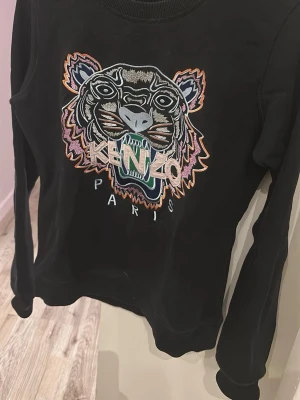 Svart Kenzo hoodie med tigerbroderi - Cool svart hoodie från Kenzo med stort broderat tigerhuvud och logga på bröstet. Färgglada detaljer i lila, orange, blå och grön. Klassisk passform och ribbade muddar. Perfekt för dig som vill sticka ut med ett ikoniskt plagg.