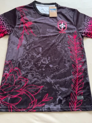 Nike Brasil sporttröja svart/rosa - Unik sporttröja från Nike med Brasil-logga. Svart bas med rosa detaljer, blommönster och leopardmönstrade ärmar. Tillverkad i lätt och ventilerande material, perfekt för träning eller match.