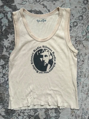 Beige ribbad linnetopp Radio Silence - Cool beige ribbad linnetopp från Brandy Melville med tryck 'Radio Silence' i svart på bröstet. Toppen är ärmlös och har en croppad passform med rå kant nedtill. Perfekt för dig som gillar en avslappnad och trendig stil. Onesize men size S