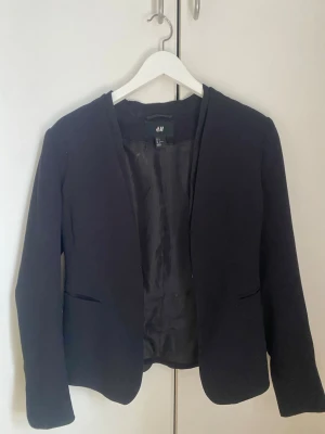 Svart kavaj från H&M - Stilren svart kavaj från H&M med öppen front och två fickor framtill. Kavajen har en klassisk, figurnära passform och är helfodrad för extra komfort. Perfekt att slänga över en t-shirt eller skjorta för en snygg look. 