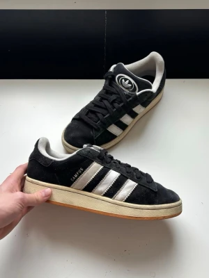 Adidas Campus svarta sneakers - Klassiska Adidas Campus sneakers i mycket bra skick. Går lätt att få dom ännu renare med en snabb tvätt. svart mocka med vita läderdetaljer och de ikoniska tre ränderna på sidan. Storlek 42