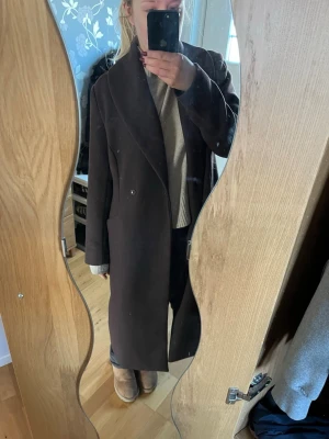 Mörkbrun lång kappa med bred krage - Säljer en mörkbrun lång kappa med bred krage och dubbelknäppning. Kappan har en rak och oversize passform, perfekt för att skapa en cool och avslappnad look. Materialet ser ut att vara ull eller ullblandning, vilket gör den varm och stilren.