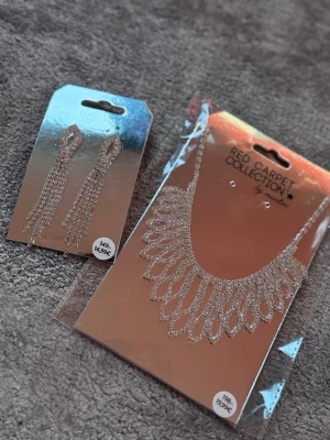 Halsband & örhängen Red Carpet - Säljer ett glittrande halsband och matchande långa örhängen från Red Carpet Collection. Båda är silvriga med massor av små strass-stenar som ger en lyxig känsla. Perfekt för dig som vill sticka ut med extra bling. Materialet är metall och strass.