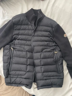 Svart pufferjacka från Moncler - Snygg svart pufferjacka från Moncler med quiltad front och ärmar i mjukt tyg. Jackan har två dragkedjeförsedda fickor och hög krage. Fylld med vitt gåsdun för extra värme. Klassisk Moncler-logga på ärmen och stilren design.