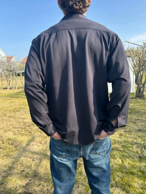 Svart långärmad skjorta - Säljer en svart långärmad skjorta med klassisk krage och knäppning vid ärmsluten. Skjortan har en avslappnad passform och är perfekt att styla med jeans. Materialet ser ut att vara bomull eller en bomullsblandning.   Jag säljer pga att den är lite lång på mig 