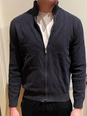 Mörkblå ziptröja med hög krage - Snygg mörkblå ziptröja med hög krage och hel dragkedja framtill. Tröjan har långa ärmar och ribbade muddar vid ärmslut och nederkant. Perfekt att slänga på sig över en t-shirt för en clean och enkel look. Dragkedjan har lossnat lite längst blir lite svårare att knäppa men det går när man har förstört hur man gör.