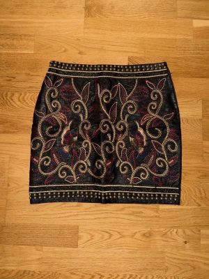 Svart kjol med broderi och gulddetaljer - Snygg svart kjol i skinnimitation med broderade mönster i guld och röda toner på framsidan. Kjolen har en rak, tight passform och dekorativa detaljer som ger en lyxig känsla. Perfekt för dig som vill sticka ut med en unik och trendig look.