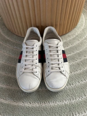 Gucci vita sneakers med röd/blå rand - Snygga vita sneakers från Gucci i läder med klassiska röd och marinblå ränder på sidorna. Lågt skaft och svart detalj vid hälen. Perfekta för dig som gillar stilrena och lyxiga skor med ikonisk design.
