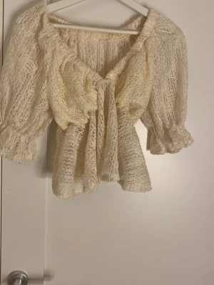 Krämvit spetsblus med volanger - Söt krämvit blus i spets med volanger och knytning framtill. Blusen har offshoulder-modell och puffiga trekvartsärmar med resår vid ärmslut. Materialet är genombrutet och ger en romantisk känsla. Perfekt för dig som gillar en bohemisk och trendig stil.