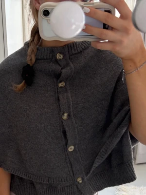 Grå stickad cape med knappar - Mysig grå stickad cape med ribbad krage och knäppning framtill med sex knappar. Capen har en avslappnad passform och breda ribbade kanter. Perfekt att slänga över en tröja för extra värme. Materialet känns mjukt och följsamt.