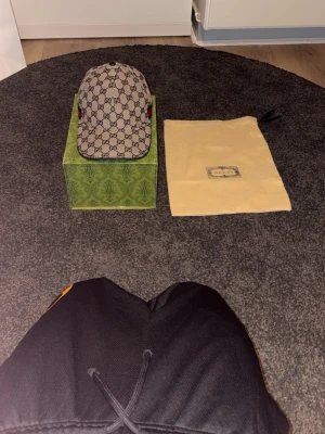 Gucci keps med GG-mönster - Snygg keps från Gucci med klassiskt GG-monogram i beige och brunt. Kepsen har böjd skärm och detaljer i rött och grönt på sidorna. Kommer med originalförpackning och dustbag. Perfekt för dig som vill sticka ut med en lyxig accessoar.