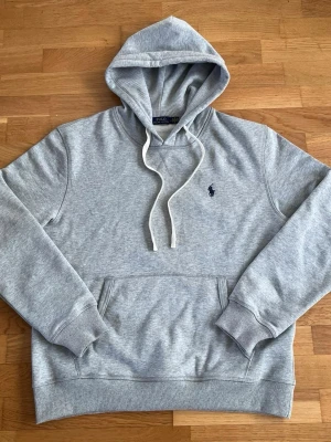 Grå hoodie från Polo Ralph Lauren - Snygg grå hoodie från Polo Ralph Lauren med klassisk huva och vita dragsnören. Har en känguruficka framtill och den ikoniska mörkblå loggan broderad på bröstet. Tillverkad i mjuk bomullsmix som känns skön mot huden. Perfekt för en chill och stilren look.