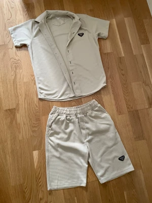 Prada loungewear set - Snyggt beige loungewear set Prada med kortärmad skjorta och matchande shorts. Setet har diskret rutmönster, knappstängning på skjortan och elastisk midja på shortsen. Båda delarna har Pradas ikoniska trekantslogga på bröstet och benet.