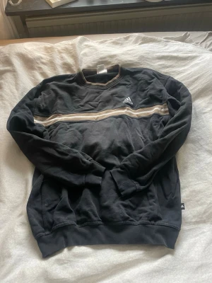 Svart sweatshirt från Adidas vintage  - Snygg svart sweatshirt från Adidas med beige och vit rand över bröstet. Tröjan har rund halsringning med beige kant och klassisk Adidas-logga på bröstet. Perfekt för en sportig och avslappnad stil. Har defekter där av säljer jag till bra pris!