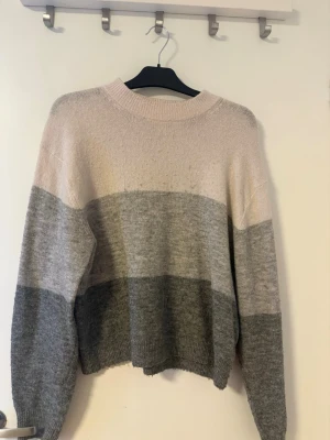 Randig stickad tröja i grå toner - Mysig stickad tröja med breda horisontella ränder i vit, ljusgrå och mörkgrå. Tröjan har en relaxed passform, rund halsringning och långa ärmar.