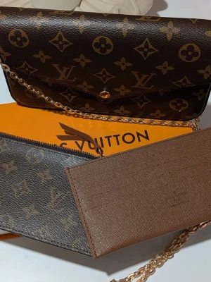 Iouis Vuitton handväska  - Väska/Kuvertväska: En rektangulär väska med ett spetsigt lock (envelop-stil) som stängs med en guldfärgad tryckknapp. Den är klädd i ett mörkt brunt mönster med geometriska former och blomliknande symboler. Blixtlåsfodral: Ett platt fodral i samma mönstrade canvas som väskan, försett med en mörk dragkedja längs ovansidan och en liten dragkläpp i läder. Korthållare: Ett platt tillbehör i ljusbrunt, texturerat läder med flera horisontella fack för kort. I mitten syns en diskret präglad logotyp. Fö