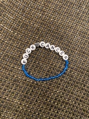 Blå pärlarmband med budskap - Ett armband med blå små pärlor och vita bokstavspärlor som stavar ut 'FUCK CANCER'. Mellan orden finns en silverfärgad hjärtformad pärla. Perfekt för att visa stöd och styrka.