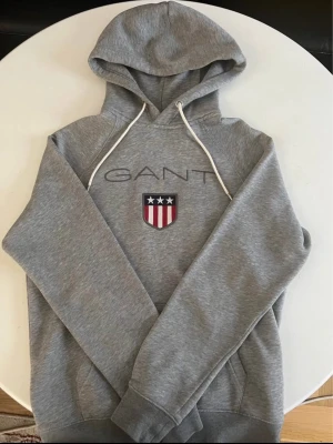Grå hoodie från GANT - Snygg grå hoodie från GANT med klassisk logga och sköld med stjärnor och ränder på bröstet. Hoodien har vit dragsko i huvan och mjukt material, perfekt för en avslappnad stil. Rak passform och ribbade muddar vid ärmar och nederkant. Priset kan diskuteras!!