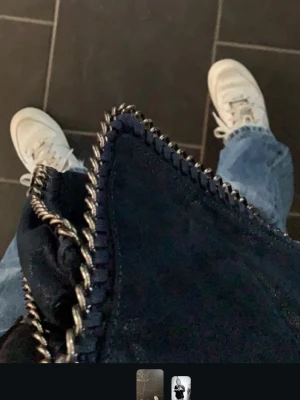 Mörkblå axelväska med kedjedetaljer - Snygg mörkblå axelväska i mockaliknande material med kraftiga silverfärgade kedjor runt kanterna och som axelrem. Väskan har en fyrkantig form och cool, trendig design som passar perfekt till jeans och sneakers.